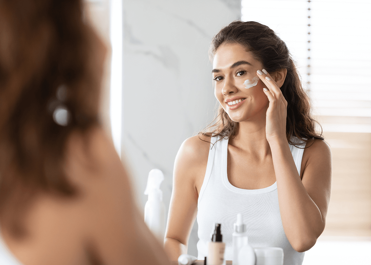 Beautiful Woman applying Moisturizer to Face in Mirror